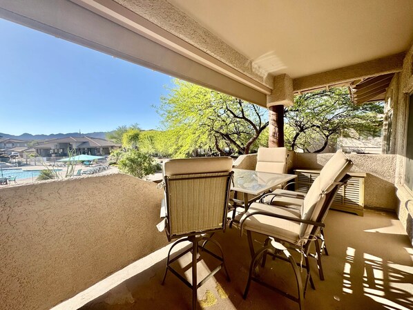 A balcony with a glass table and wicker chairs, overlooking a swimming pool and a mountainous landscape.