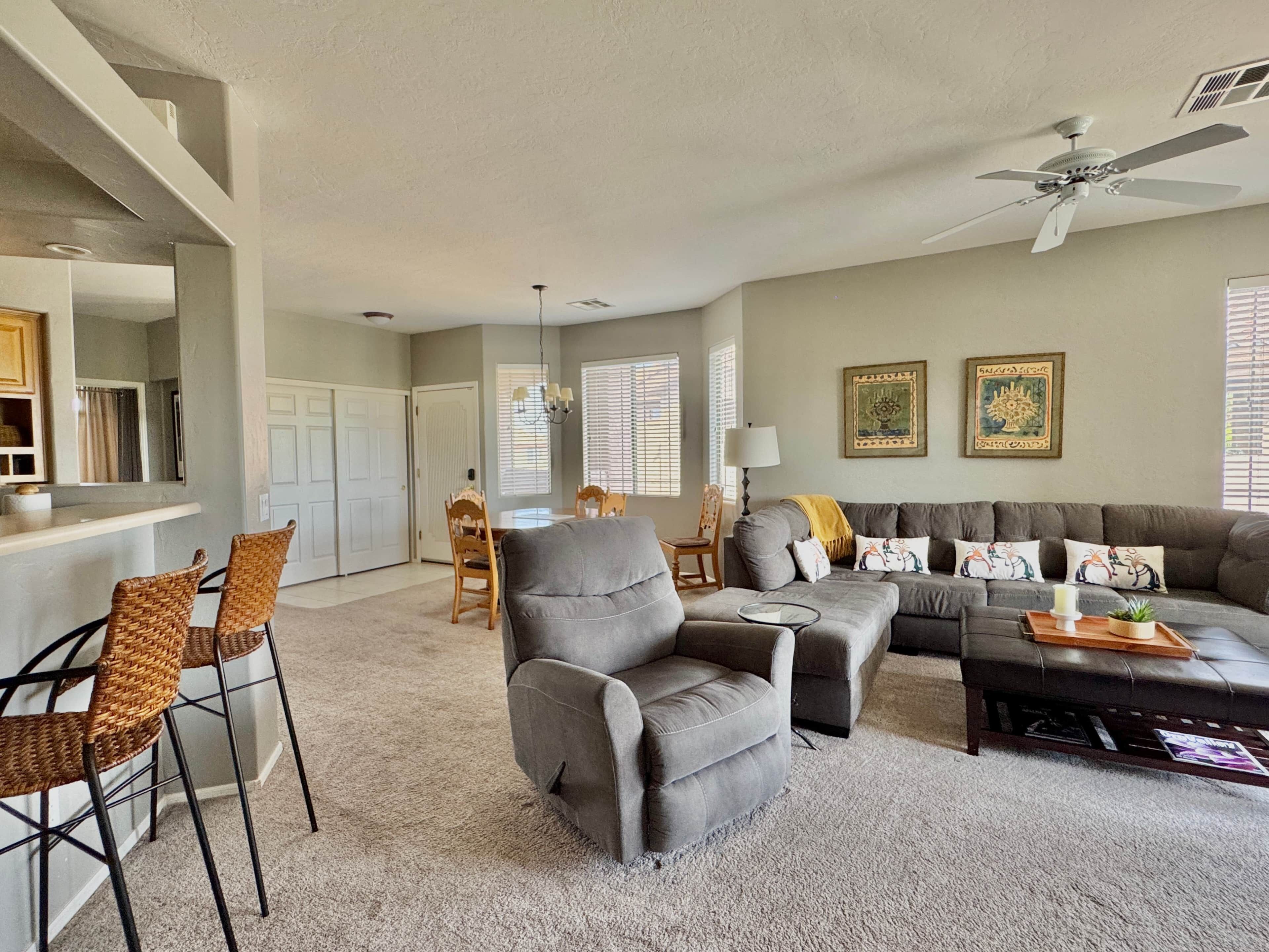 A spacious living room with a gray sofa, two armchairs, and a coffee table. There is a ceiling fan, a dining area with a table and chairs, and a kitchen area with a counter and cabinets.
