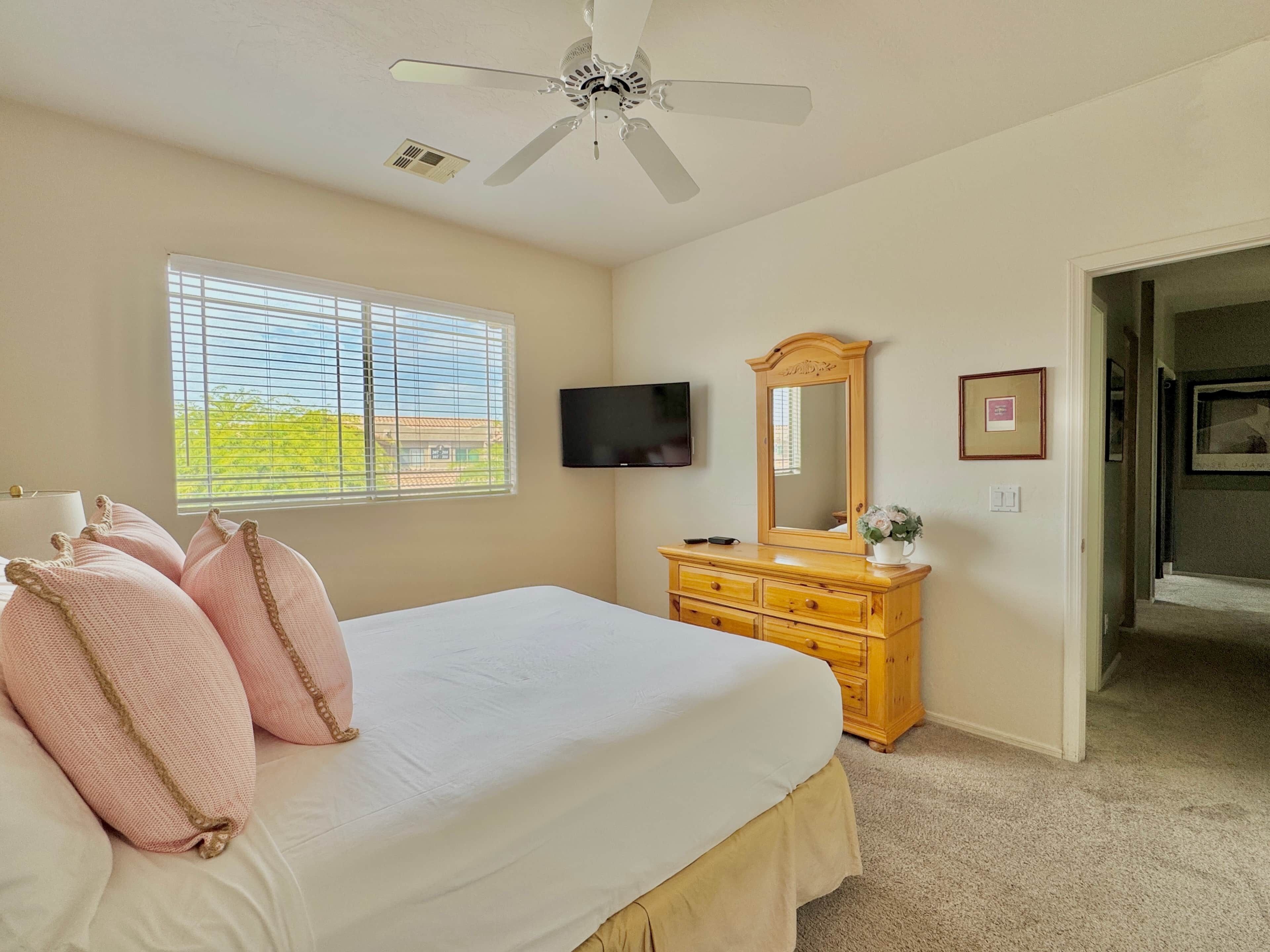 A bedroom with a bed, a dresser, a television, a window with blinds, and a ceiling fan.