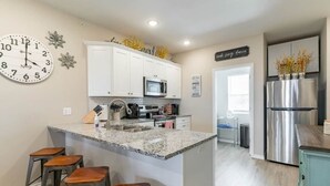 A modern kitchen with white cabinets, a granite countertop, and stainless steel appliances. There are three stools at the island and a clock on the wall.