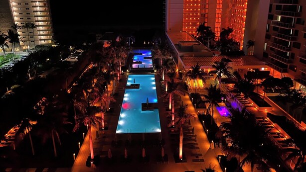 Une vue nocturne d’un hôtel avec une piscine, des palmiers et un bâtiment illuminé.