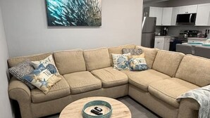 A living room with a beige sectional sofa, a round coffee table, and a painting on the wall. - Dolphin Cove | Steps from the Beach | 2BR w/Hockey/Ping-pong/Pool Table (Cocoa Beach)