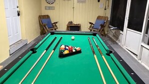 A pool table with balls, chairs, and a ceiling fan in a room with a door and window. - Dolphin Cove | Steps from the Beach | 2BR w/Hockey/Ping-pong/Pool Table (Cocoa Beach)