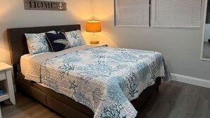 A bedroom with a bed, a ceiling fan, a bedside lamp, a sign reading "HOME", and a window with blinds. - Dolphin Cove | Steps from the Beach | 2BR w/Hockey/Ping-pong/Pool Table (Cocoa Beach)