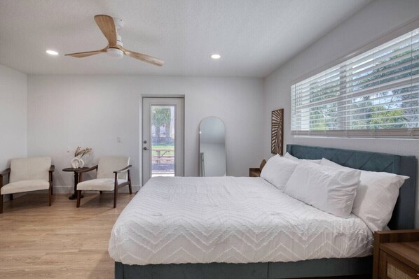 A bedroom with a bed, two chairs, a small table, a ceiling fan, and a window with blinds.
