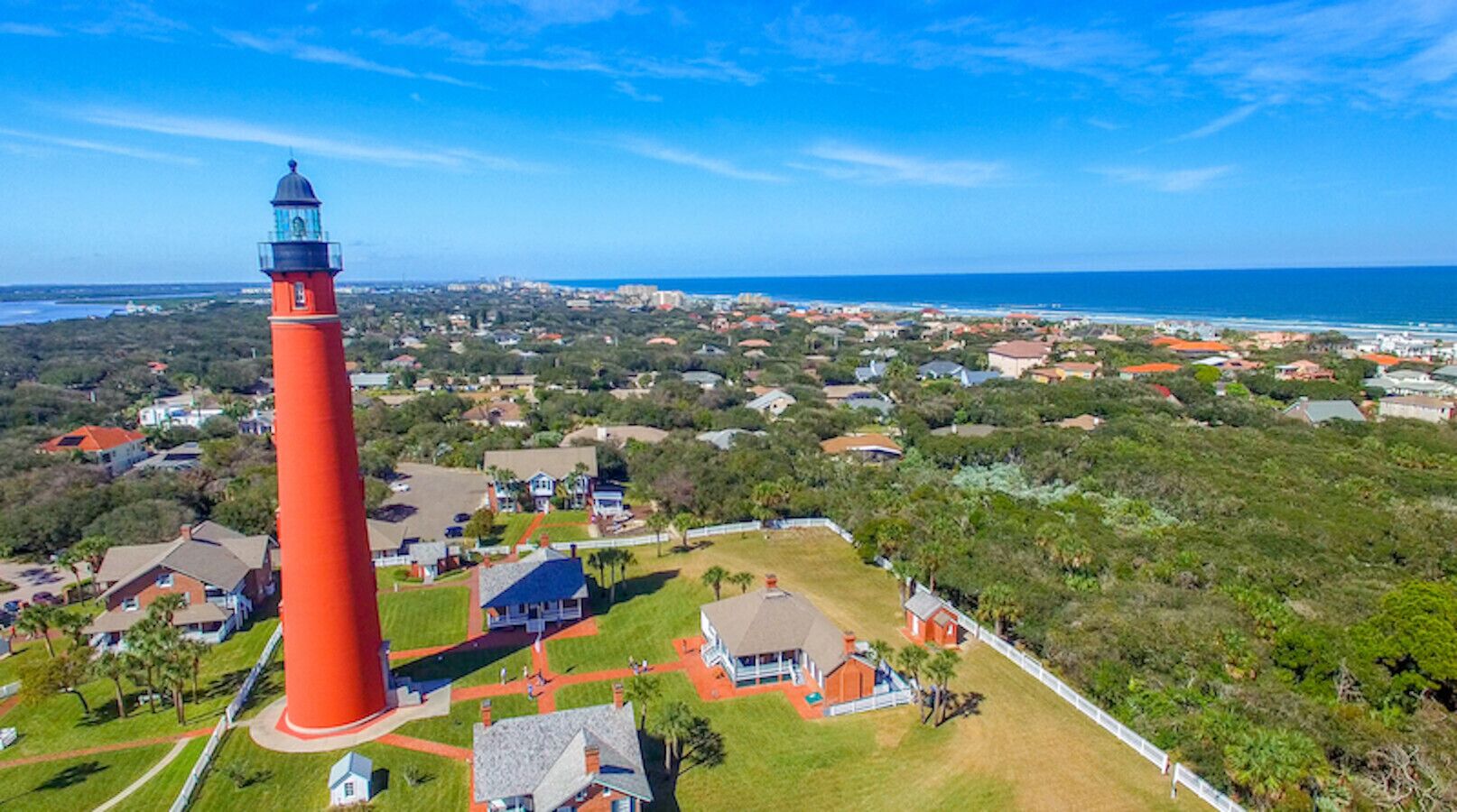 An aerial view of a coastal town with a prominent red lighthouse, residential houses, and a clear view of the ocean.