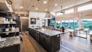 A modern kitchen with a large island, wooden ceiling, and large windows offering a mountain view.