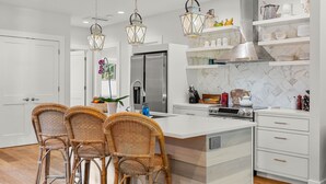 A modern kitchen with a large island, wicker chairs, white cabinetry, and hanging lanterns. - Modern Gorge Retreat • Hot Tub • Mountain Views (Cascade Locks)