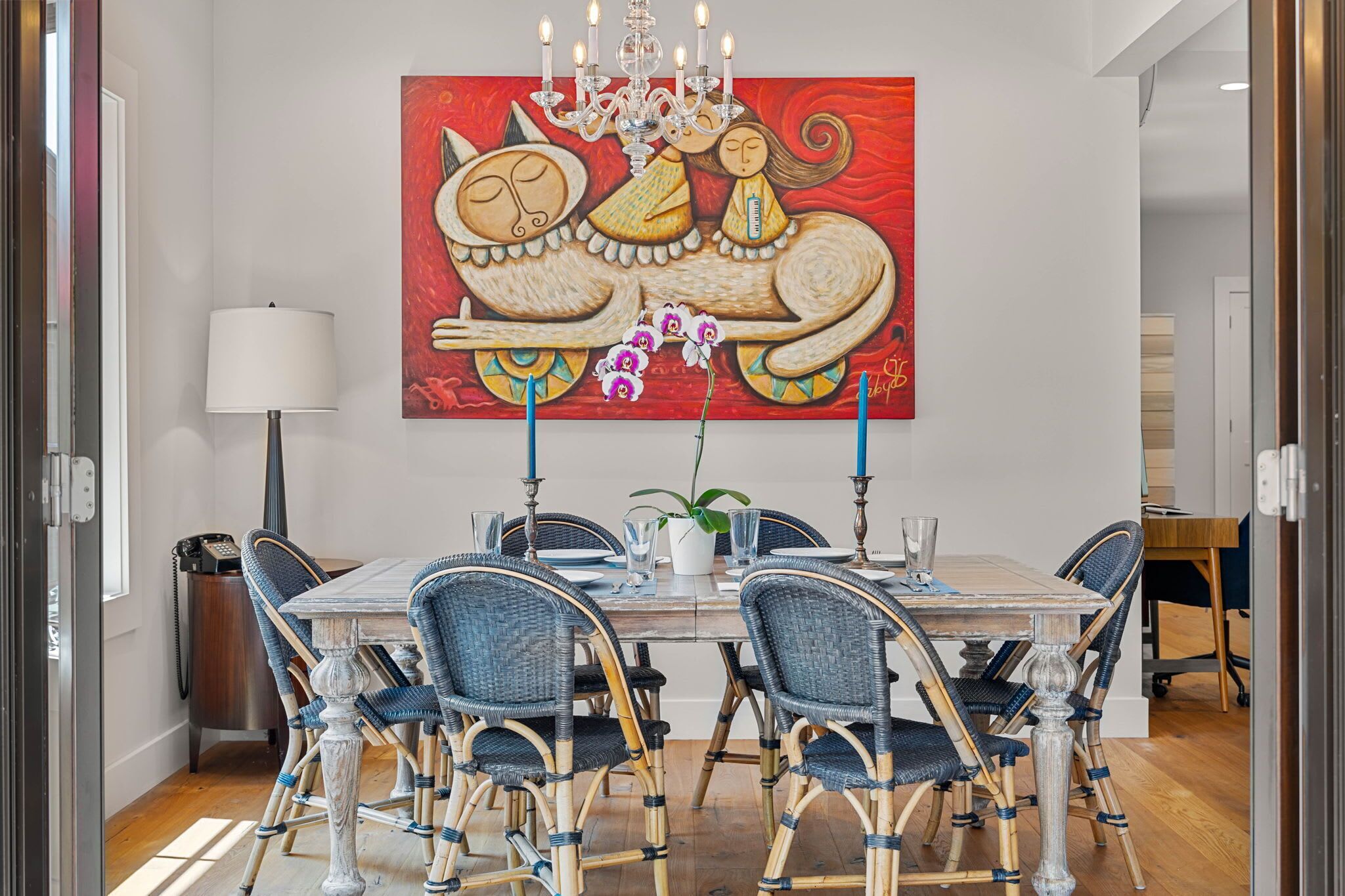 A dining area with a table set for six, surrounded by wicker chairs, a chandelier, and a large painting on the wall.