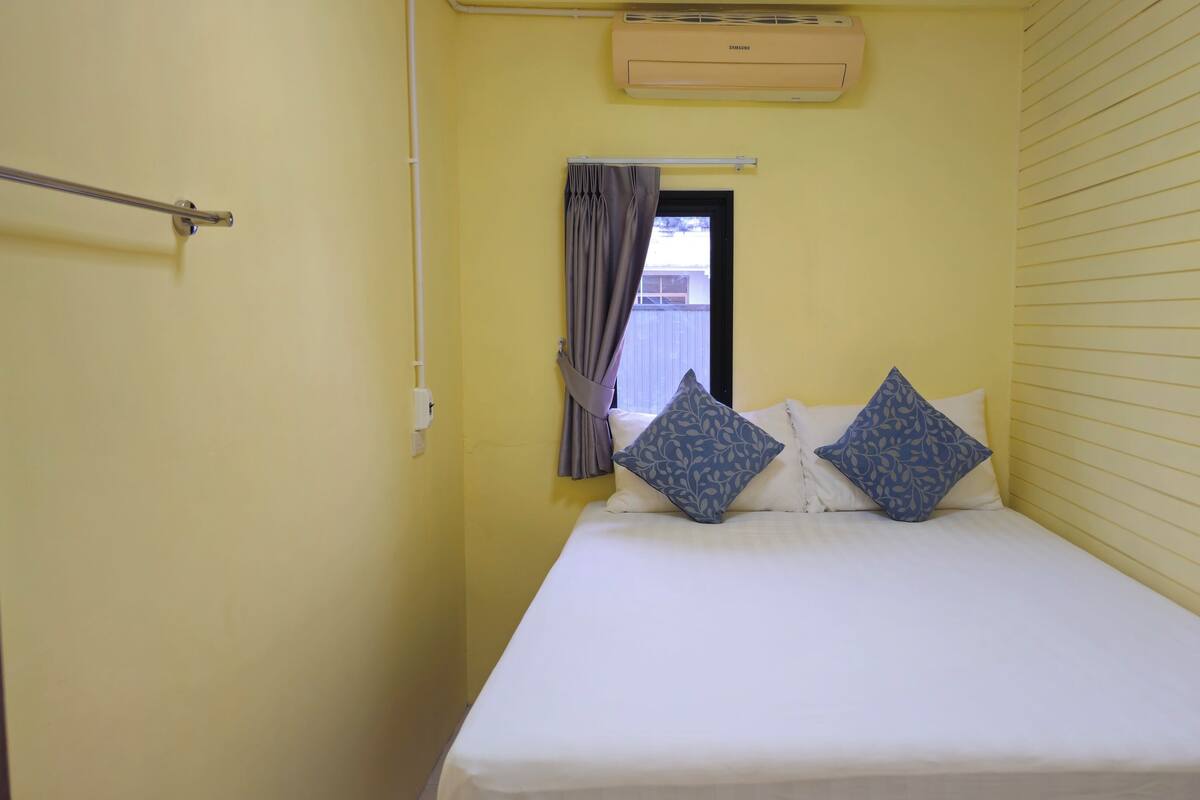 A small, narrow room with a bed, two pillows, a window with curtains, and an air conditioning unit above the window.