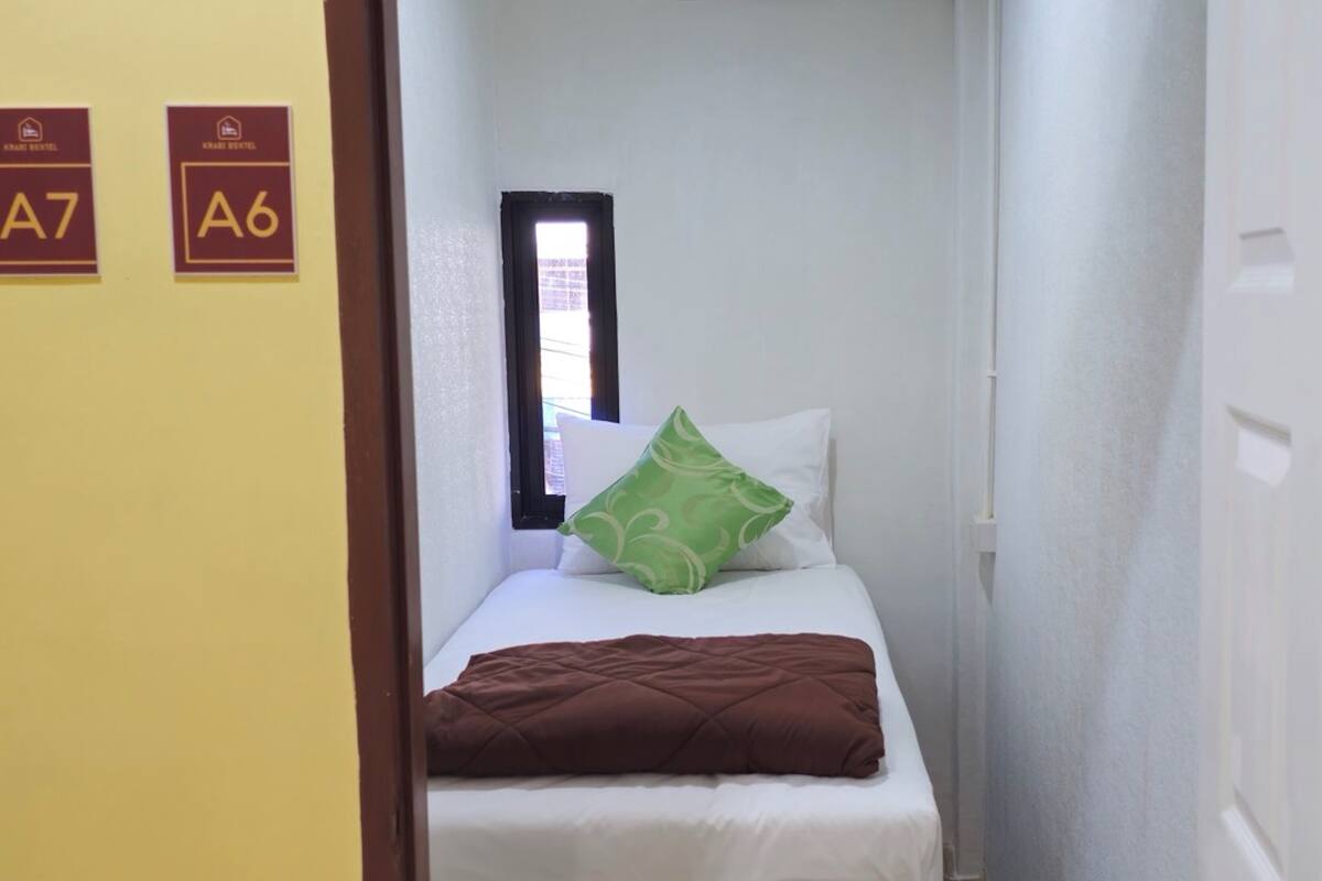 A narrow hotel room with a bed, a green pillow, and a brown blanket. There are two signs on the wall indicating room numbers A7 and A6.