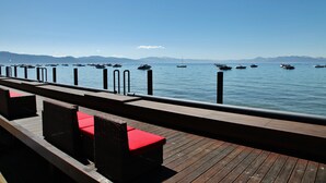 A wooden deck with wicker chairs and red cushions, overlooking a calm sea with boats and distant mountains. - Tahoe Vista Inn Suite 7 (Tahoe Vista)