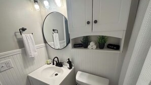 Et badeværelse med en hvid vask, vandhane i sort farve og et toilet. Der er et spejl over vasken samt en hylde med planter og toiletartikler.