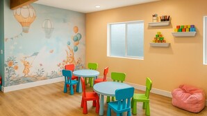 Ein Kinderspielzimmer mit bunten Stühlen, einem runden Tisch, einer Wandmalerei mit Heißluftballons und einem Fenster mit Jalousien.