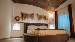 A circular bed with a headboard, a wall-mounted shelf with decorative items, and a window with a view of buildings. - Suite with private spa in the heart of Palermo✨️ (Palermo)