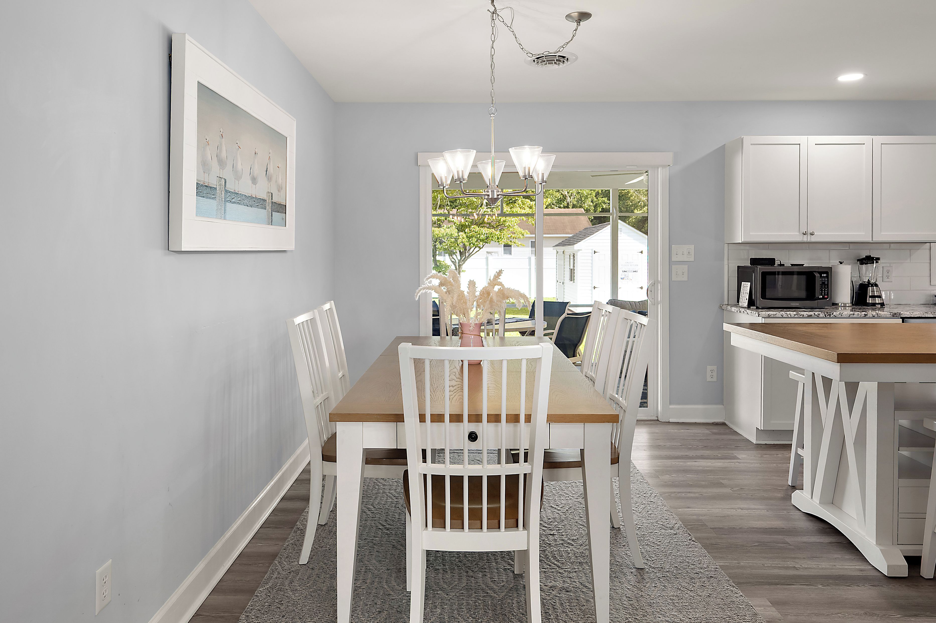 A modern kitchen with white cabinetry, a dining area with a wooden table and white chairs, and a view through the door to the outside.