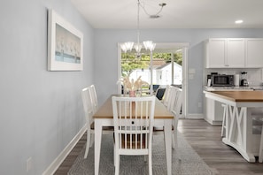 Een moderne keuken met witte kasten, een eethoek met een houten tafel en witte stoelen, en uitzicht door de deur naar buiten.
