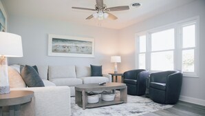 A living room with a grey sofa, a coffee table, a black armchair, a ceiling fan, and a painting of birds on the wall. - 4BR Beach House Sleeps 12/Screened Porch/Parking for 6 cars /Rehoboth (Rehoboth Beach)