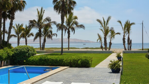 A pool area with lounge chairs, palm trees, and a view of the sea. - Casa Paracas by Wynwood House (Paracas)