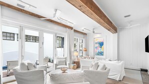 A spacious living room with white furniture, large windows, and a wooden ceiling beam.