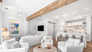A modern living room with white sofas, a coffee table, and a kitchen area with white cabinets and a wooden ceiling beam.