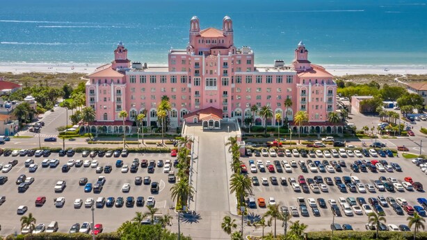 Un edificio grande de color rosa con una cúpula central y dos torres, rodeado por un estacionamiento y palmeras, cerca de una playa.