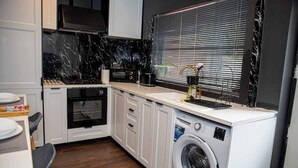 Una cocina compacta con armarios blancos, horno empotrado, lavadora y ventana con persianas.