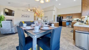 Een moderne keuken met roestvrijstalen apparatuur, een eethoek met een houten tafel en blauwe stoelen, en een woonkamer met een grijze bank en een flatscreen-televisie.