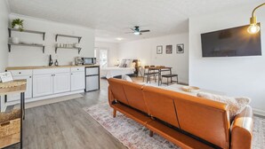 A modern kitchen with white cabinets, a dining area with a brown leather sofa, and a flat-screen TV mounted on the wall.