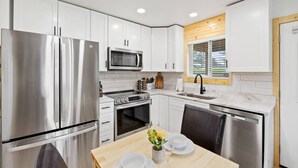 A modern kitchen with white cabinets, stainless steel appliances, and a wooden dining table set for two. - Custer Love Shack Getaway (Custer)