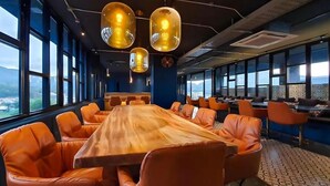 A conference room with a long wooden table surrounded by orange chairs, large windows offering a view of the mountains, and hanging pendant lights. - Infinity Sky Pool Walk to BeachC182 (Kamala)