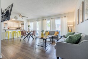 Ein modernes Wohnzimmer mit einem grauen Sofa, einem Couchtisch und gelben Sesseln. Im Hintergrund ist eine Küche mit weißen Schränken und einer Mikrowelle zu sehen.
