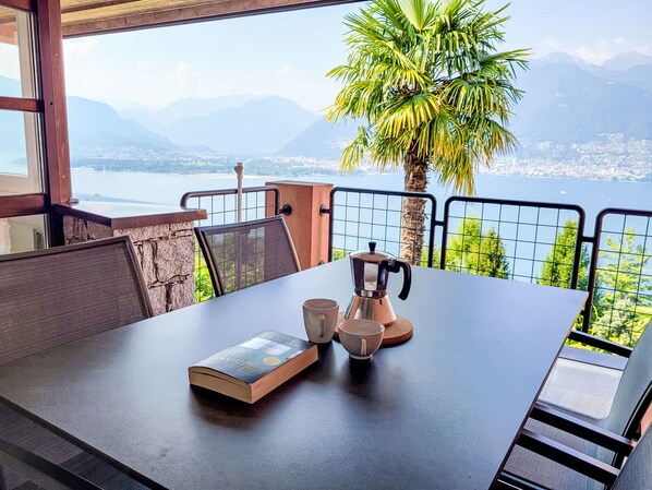 Uma mesa com um livro, duas xícaras e uma cafeteira numa varanda com vista para um lago e montanhas.