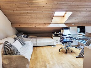 Quarto no estilo loft com teto de madeira, claraboia, cama com almofadas estampadas, escrivaninha com computador e uma cadeira.