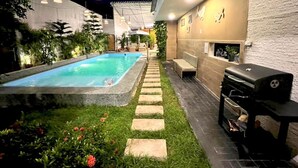 Un patio trasero con piscina, escalones de piedra, una parrilla y luces exteriores.