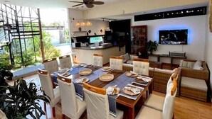 Un comedor moderno con una mesa grande preparada para seis comensales, rodeada de sillas acolchadas. Se ve una cocina con un televisor de pantalla plana al fondo.