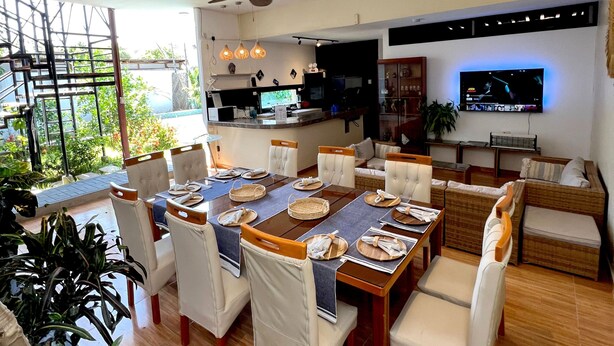 A modern dining area with a large table set for six, surrounded by white upholstered chairs. A kitchen with a black countertop and a television mounted above the counter is visible in the background.