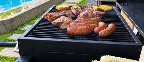 A barbecue grill with various meats and vegetables, a pool in the background, and outdoor furniture.