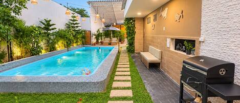 A swimming pool with a tiled edge, surrounded by a garden with green grass and red flowers. There is a barbecue grill and outdoor seating area with a bench.