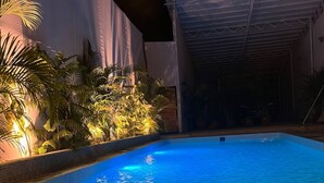 An indoor swimming pool with tiled edges, surrounded by potted plants and a covered area in the background.