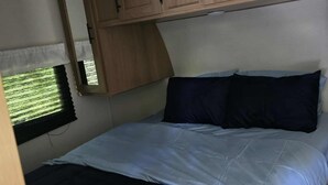 A compact RV interior with a bed, a window with blinds, and wooden cabinets.