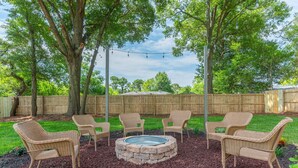 An outdoor seating area with wicker chairs and a stone fire pit surrounded by mulch, set against a wooden fence and trees. - THE GARDEN HOUSE (Fort Walton Beach)