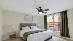 Une chambre à coucher comprenant un lit, une table de chevet, une lampe, un ventilateur de plafond, une fenêtre avec des rideaux et une vue sur les bâtiments à l’extérieur.
