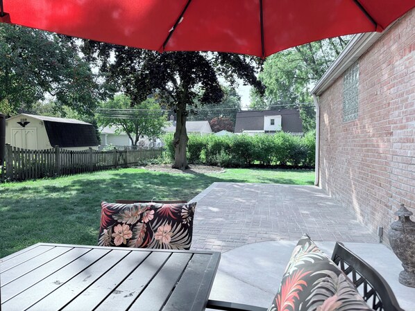 Un patio con una mesa de madera y un paraguas rojo, una pared de ladrillo y una valla de madera al fondo.