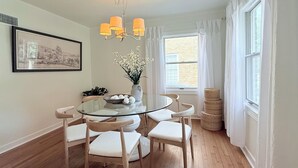 Un coin repas avec une table en verre entourée de chaises en bois, un lustre et une fenêtre avec des rideaux.