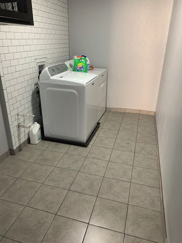 A laundry room with a front-loading washing machine, a detergent bottle, and a white trash bin.