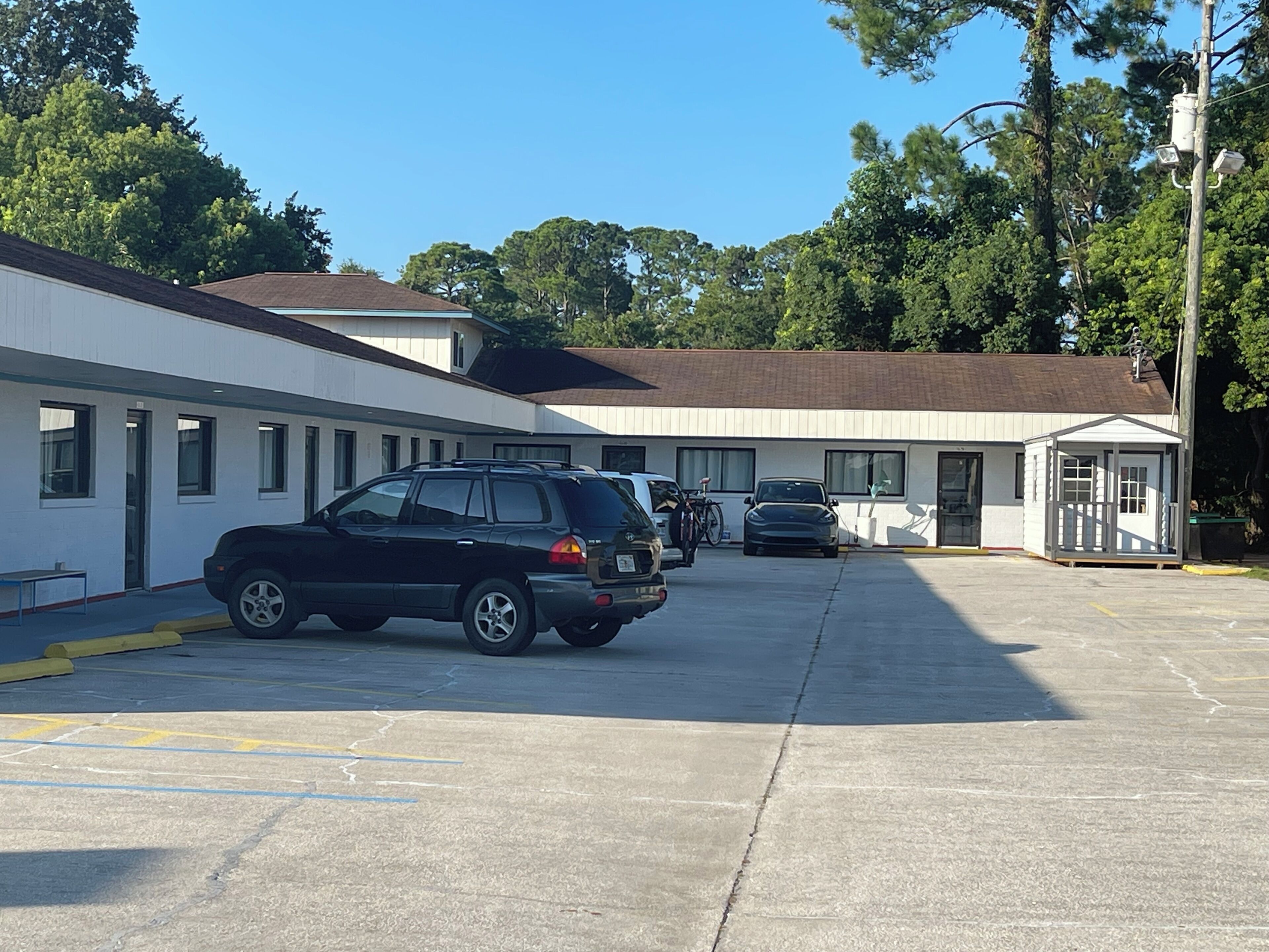 A parking lot with several cars, a small building with a porch, and trees in the background.