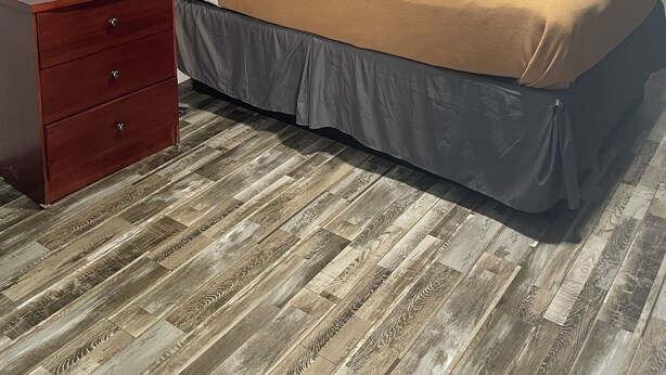 Cama bem arrumada com cabeceira de madeira, mesa de cabeceira com gavetas e luminária fixada na parede.