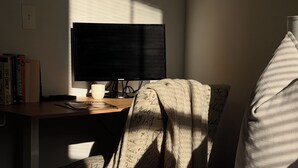 Een kamer met een bureau, een beeldscherm en een stoel met een deken. Er staan boeken op het bureau en een kussen op het bed.