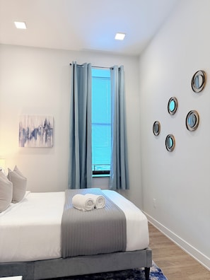 Une chambre à coucher moderne avec un lit, une fenêtre ornée de rideaux bleus, un miroir fixé au mur et un tableau représentant un paysage urbain.